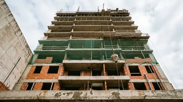 Prédio residencial em construção visto da calçada, com estrutura de concreto, alvenaria de bloco cerâmico e tela de proteção na fachada.